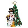 A jolly snowman with gift and broom in hand stands in front of a holiday decorated tree.