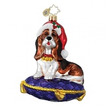 A cute basset hound puppy, perched on a glittering tasseled pillow in a santa hat and gold accented collar.